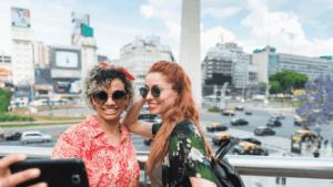 Fotografía de dos chicas haciendo un selfie, de fondo el Obelisco de Buenos Aires. 