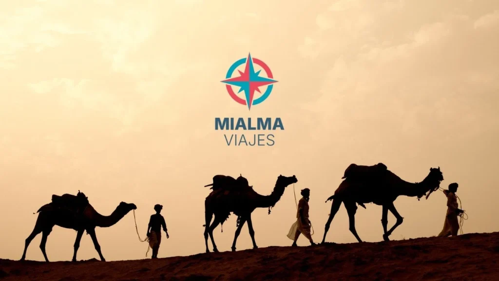 Tres camellos tirados por tres hombres en desierto. Logo de Mialma viajes en el centro de la imagen. 