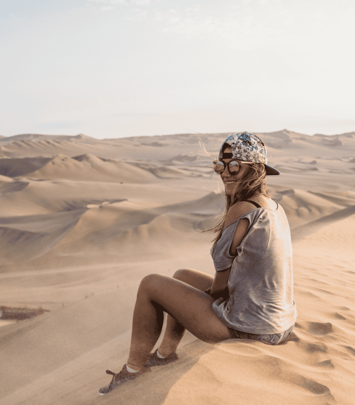 Mujer sentada en desierto con gorra y gafas. Logo de Mialma Viajes, agencia de viajes en familia.